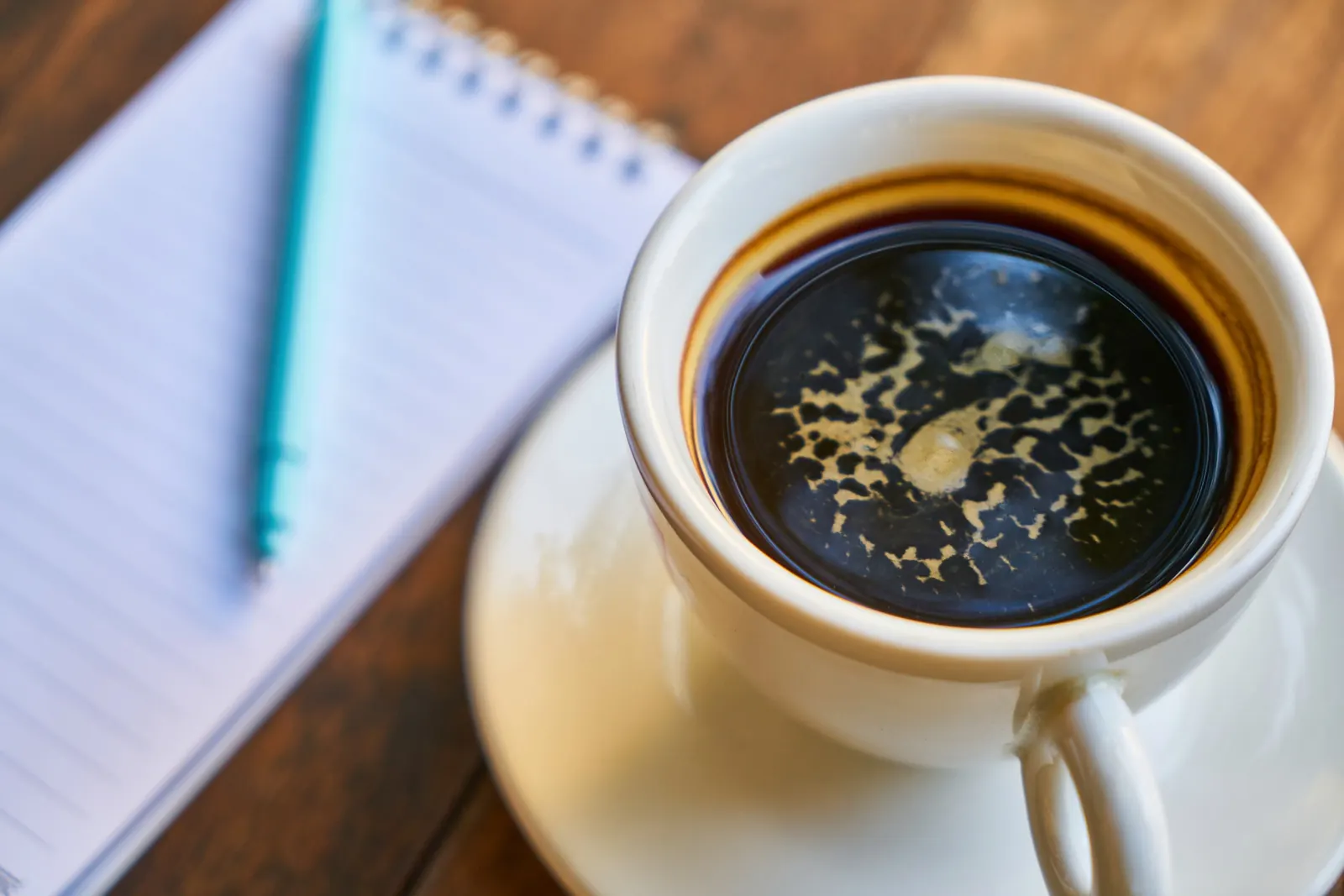 An open notebook and a pen beside a cup of coffee on a wooden desk.